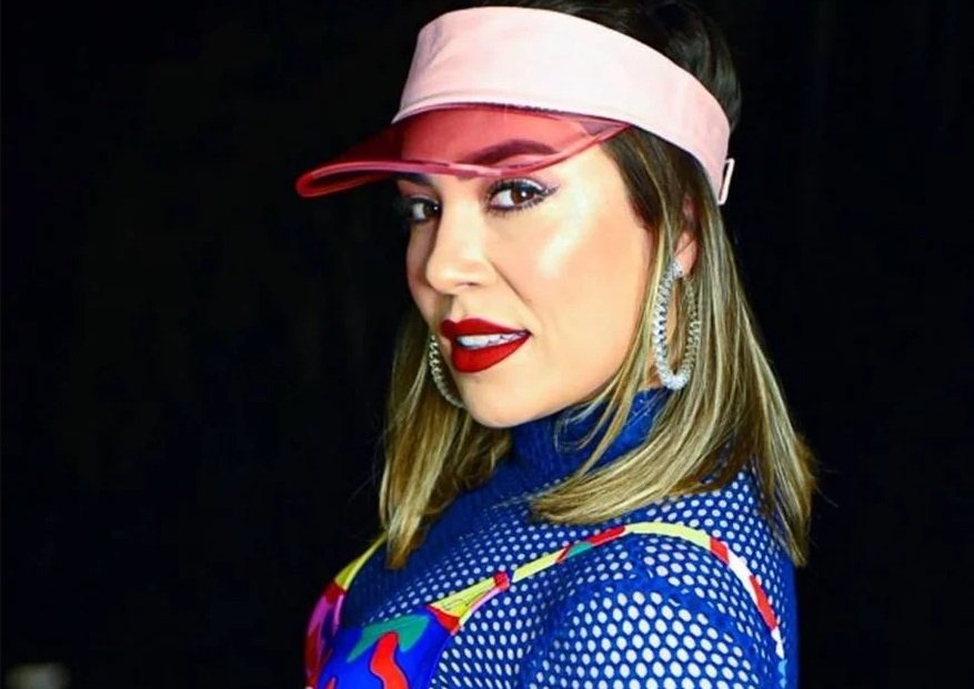 Na imagem colorida, uma mulher está posicionada no centro. Ela tem cabelos curtos e loiros, olhos castanhos e olha seriamente para a câmera