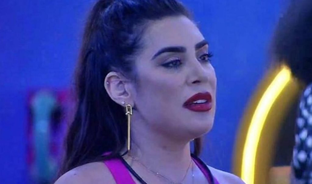 Na imagem colorida, uma mulher está posicionada no centro. Ela tem cabelos curtos e loiros, olhos castanhos e olha seriamente para a câmera