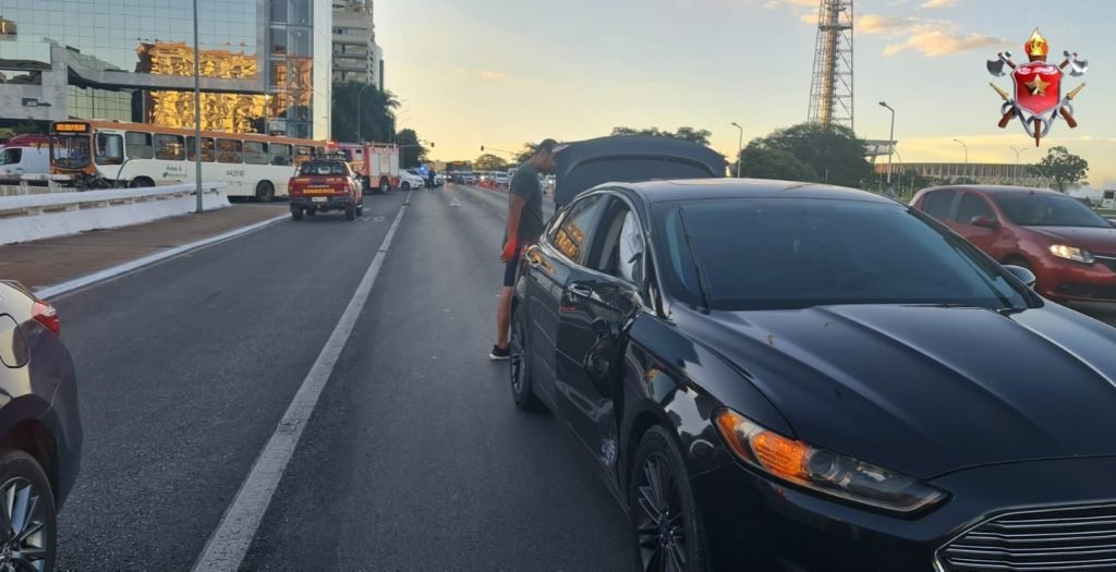 Carro preto em pista e ônibus batido em muro ao fundo