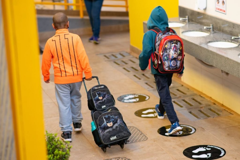 Crianças de costas, com mochilas, caminhando em escola
