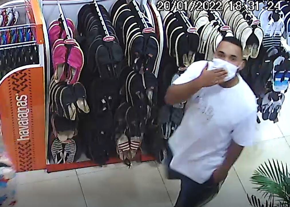 Homem com camisa branca e com a mão em máscara. Ele está dentro de loja de chinelos