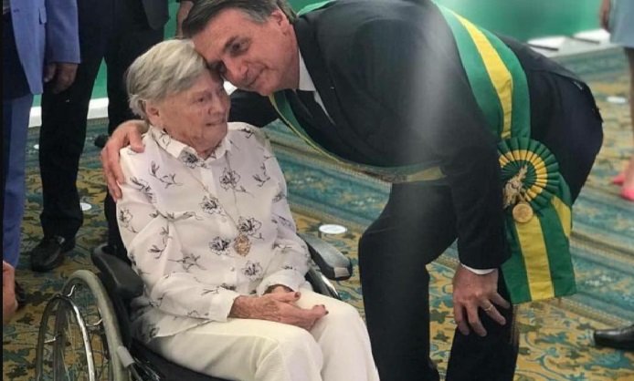 Na imagem colorida, um homem e uma mulher estão posicionados no centro. Os dois têm cabelos curtos e grisalhos, o homem usa terno e a senhora está sentada em uma cadeira de rodas. Ela usa roupa clara