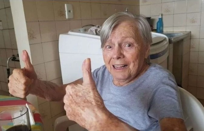 Na imagem colorida, uma senhora está posicionada no centro. Ela tem cabelos brancos e curtos, usa camiseta cinza e está fazendo sinal de joia com uma das mãos