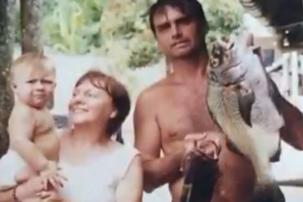 Na imagem colorida, um homem e uma mulher estão posicionados no centro. Os dois têm cabelos curtos e grisalhos. O homem usa bermuda e está sem camisa. A mulher usa roupas claras e segura uma criança no colo