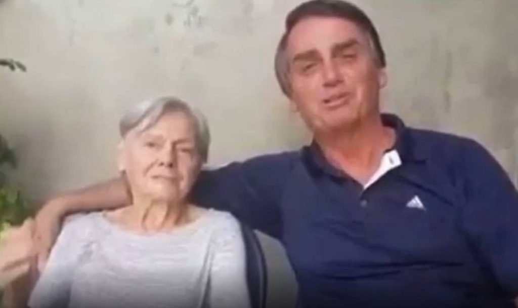 Na imagem colorida, um homem e uma mulher estão posicionados no centro. Os dois têm cabelos curtos e grisalhos, usam camiseta e bermuda e sorriem para a camera