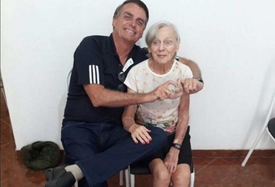 Na imagem colorida, um homem e uma mulher estão posicionados no centro. Os dois têm cabelos curtos e grisalhos, usam camiseta e bermuda e sorriem para a camera
