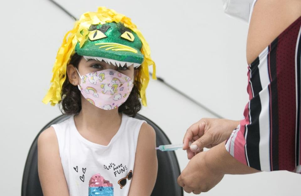Menina com chapéu de jacaré na cabeça e máscara sendo vacinada