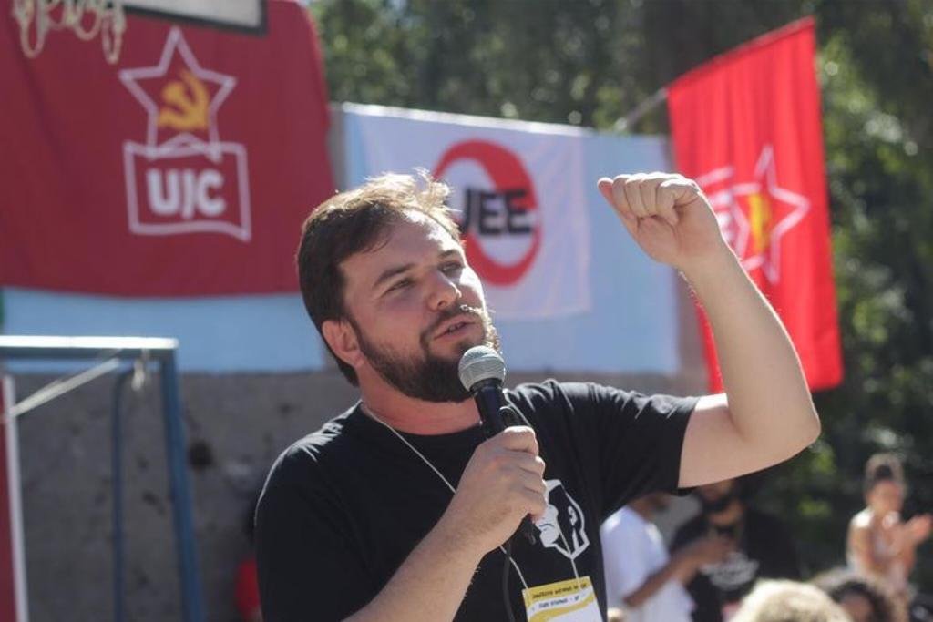 Na imagem colorida, um homem está posicionado no centro. Ele tem cabelos curtos, pretos e olha seriamente para frente