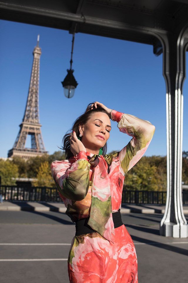 Torre Eiffel ao fundo de imagem. Mulher aparece desfilando e com os olhos fechados. Ela está com vestido em tons de verde, rosa e vermelho