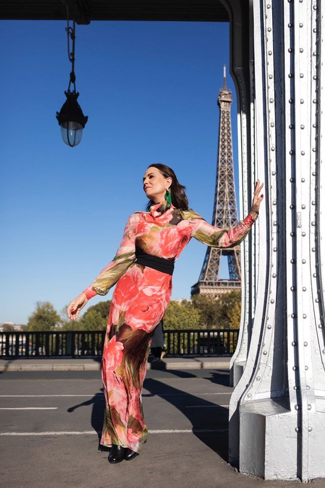 Torre Eiffel ao fundo de imagem. Mulher está escorada em uma pilastra. Ela está com vestido em tons de verde, rosa e vermelho
