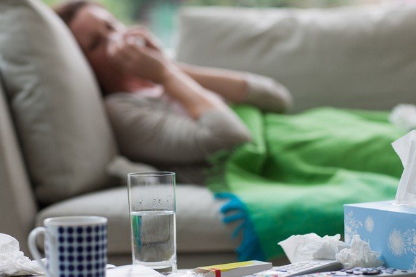 mulher doente assoa nariz em papel. Ela está deitada no sofá ao lado de medicamentos-Metrópoles