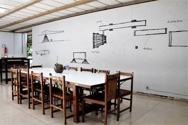 Foto mostra mesa e painel com desenhos de arquitetura ao fundo