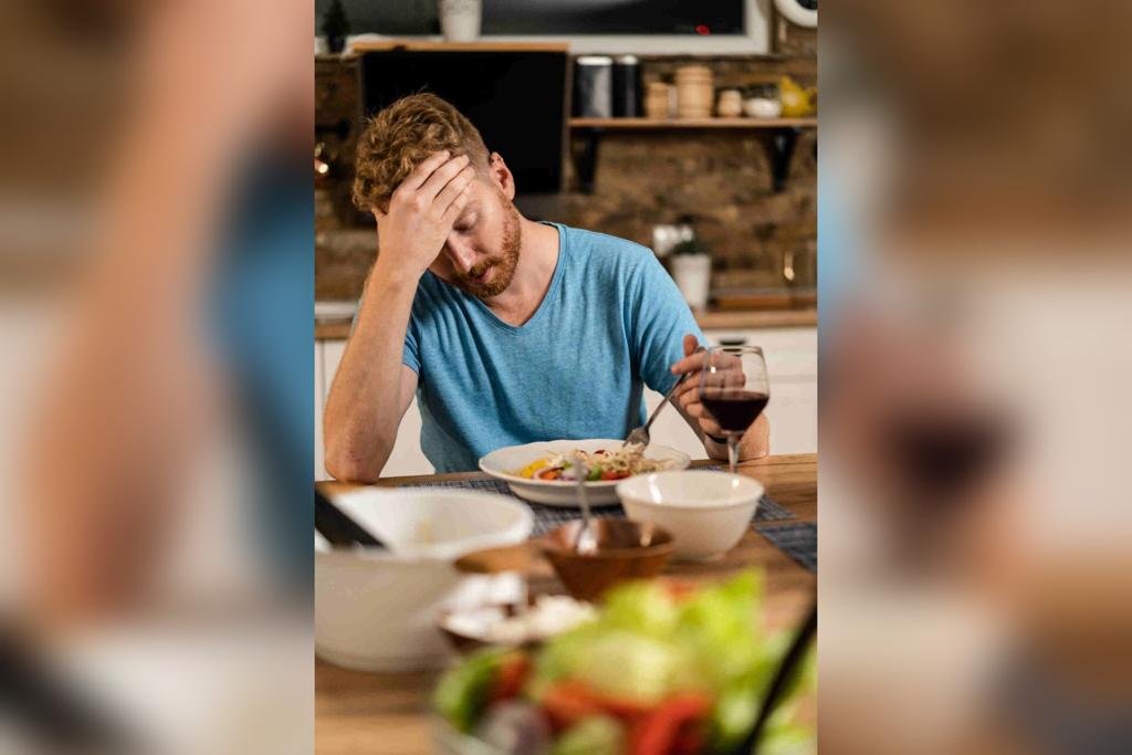 Na imagem colorida, uma pessoa está posicionada no centro. Ele está sentado em uma cadeira em frente a uma mesa com comida. Está com uma das mãos na cabeça enquanto olha para um prato com comida