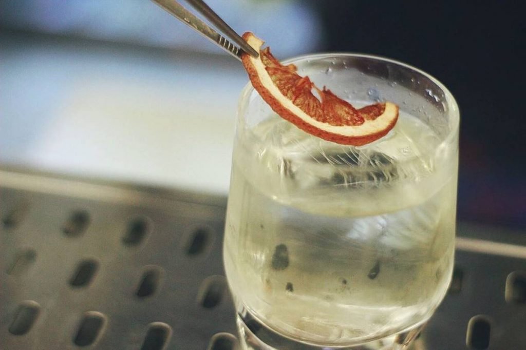 Na foto, um copo baixo com liquido transparente e uma pinsa colocando meia fatia de toranja desidratada sobre o liquido