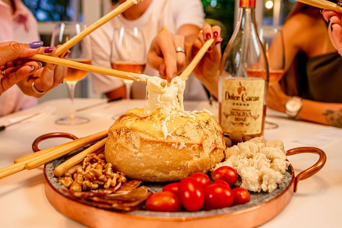 Na foto temos uma tabua de cobre com um pao italiano cheio de fondue de queijo com nozes, tomates cereja e cubos de pao ao redor e uma garrafa de vinho ao fundo, além de taças