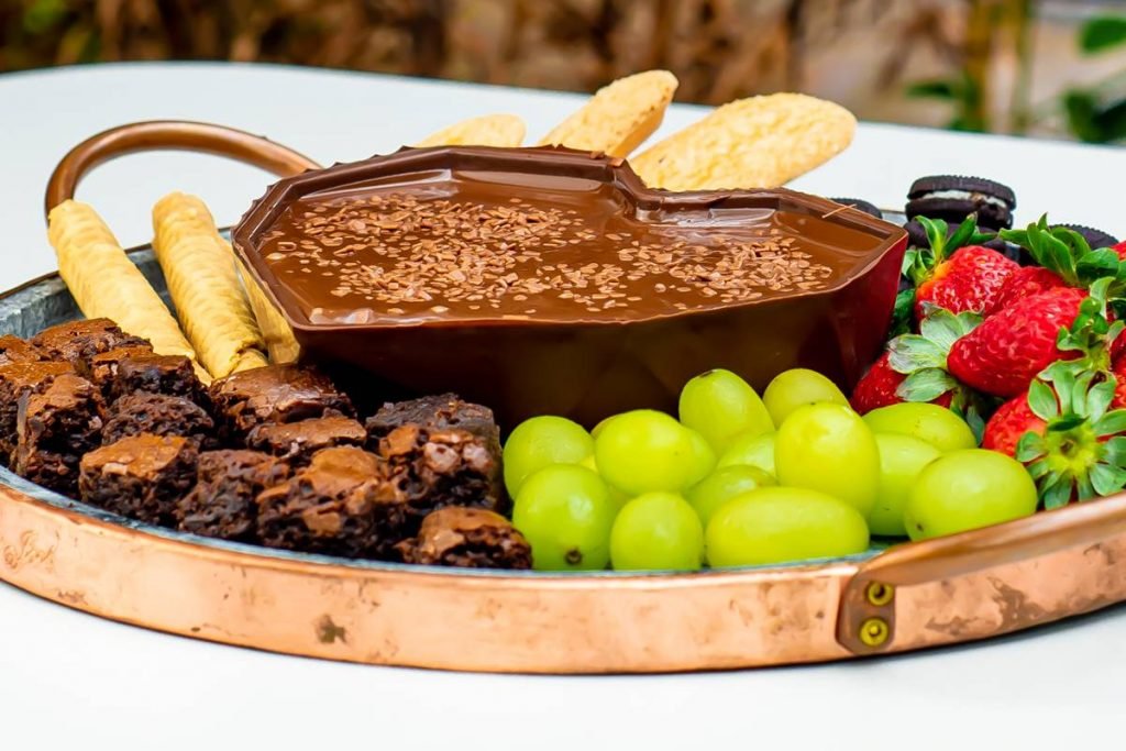 Na foto temos uma tabua de pedra com um coração de chocolate cheio de chocolate derretido e biscoitos, morangos, uvas e brownies ao redor vista de lado
