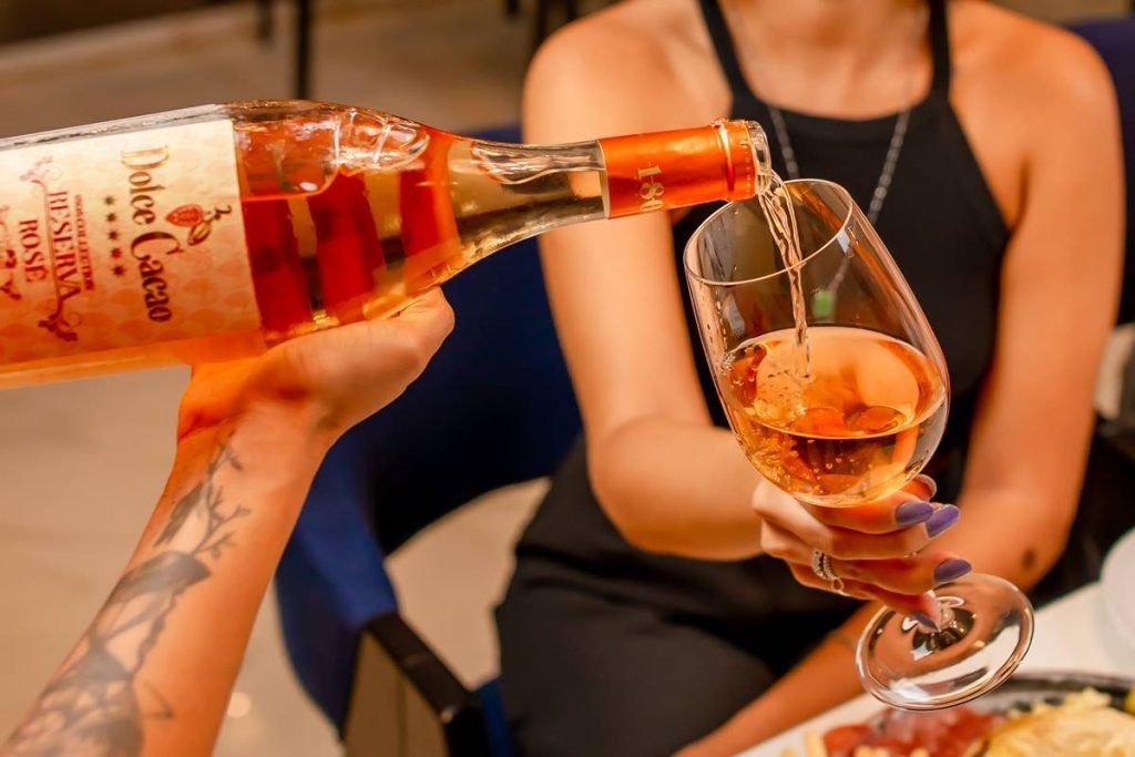 na foto temos o corpo de uma mulher segurando uma taça de vinho e outra mao colocando vinho rose na taça