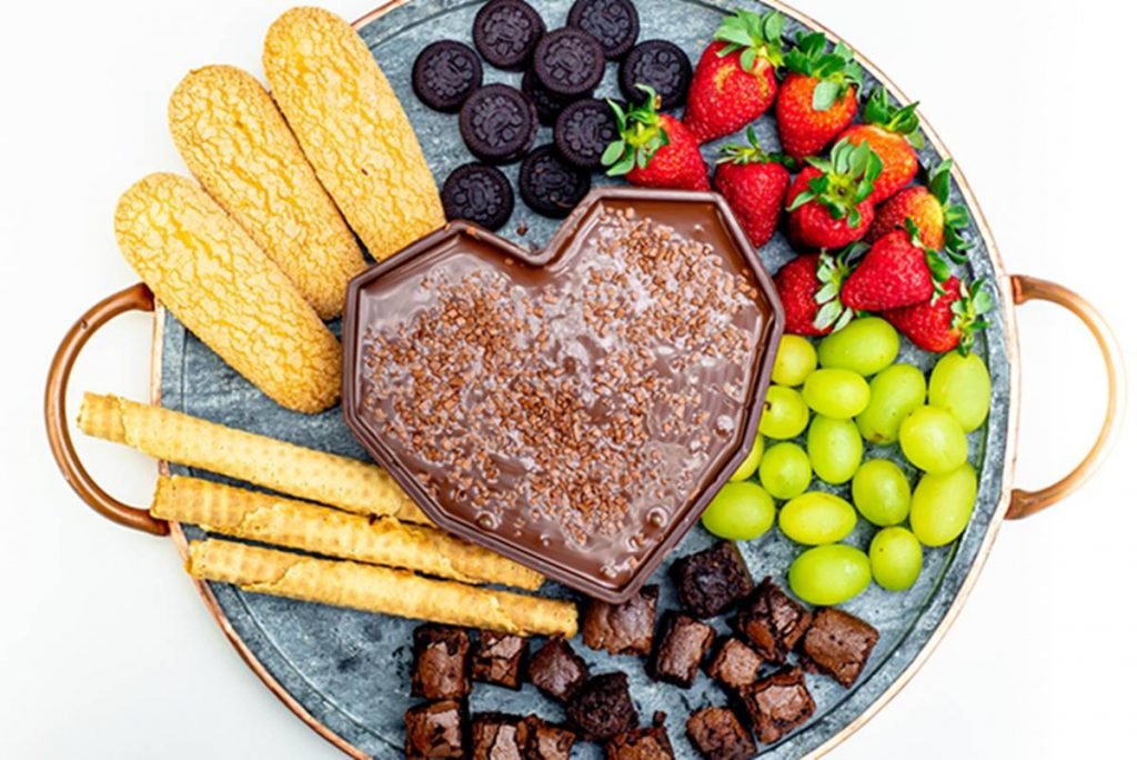 Na foto temos uma tabua de pedra com um coração de chocolate cheio de chocolate derretido e biscoitos, morangos, uvas e brownies ao redor