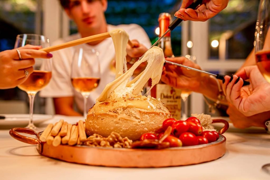 Na foto temos uma bandeja de cobre com pão italiano cheio de queijo no meio, tomate cereja, nozes e grissinis ao redor. Ao fundo vemos uma parte de uma pessoa, taças e uma garrafa de vinho