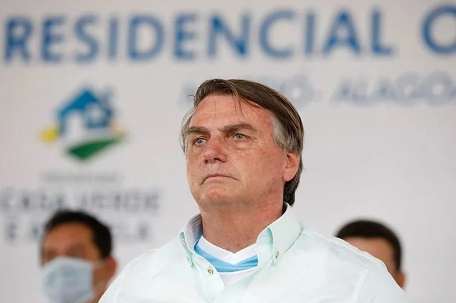 Na imagem colorida, um homem está posicionado no centro. Ele tem cabelos curtos, pele clara e olha seriamente para a frente
