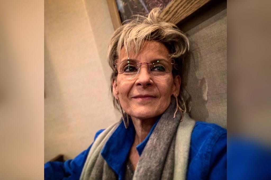 Na imagem colorida, uma mulher está posicionada no centro. Ela tem cabelos curtos e brancos, casaco azul e sorri para a camera