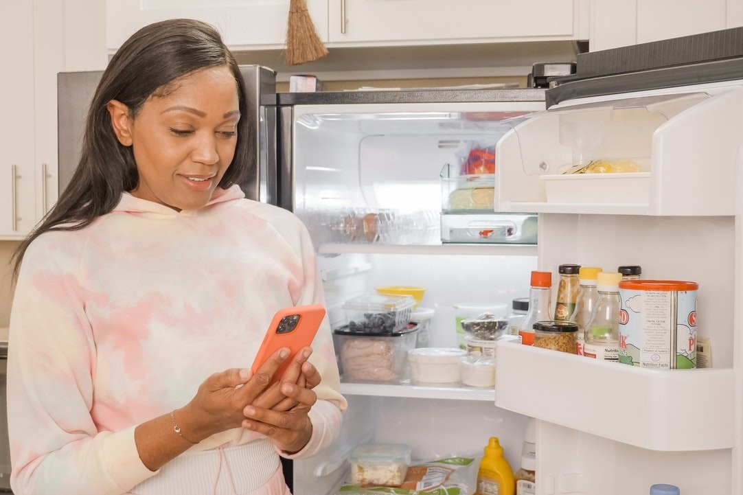 Mulher olhando o celular com geladeira aberta ao fundo