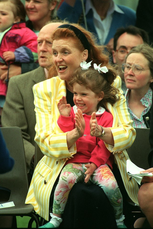 Mulher de blazer amarelo com uma menina sorridente no colo. Elas batem palmas