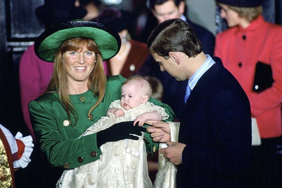 Mulher vestida com roupa verde. Ela segura uma bebê no colo. Homem de terno ao lado