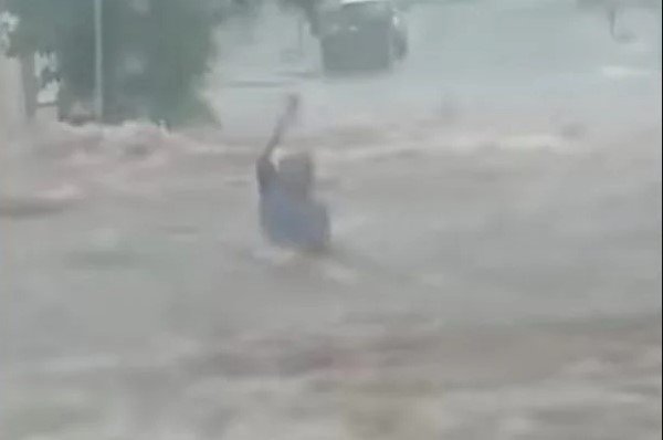 Na imagem colorida, uma pessoa fica em pé no meio de um local alagado com os braços levantados