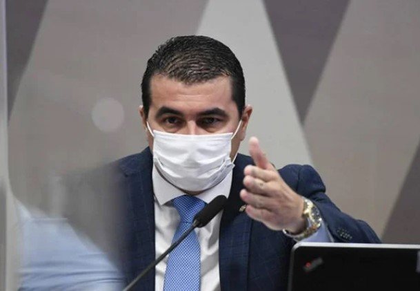 Na imagem colorida, um homem está posicionada no centro. Ele usa terno e gravata na cor preta e camiseta branca. Ele olha seriamente para a camera
