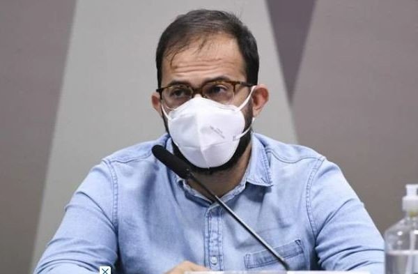Na imagem colorida, um homem está posicionada no centro. Ele usa máscara branca, camiseta clara e olha para frente