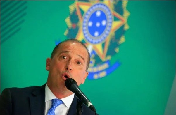 Na imagem colorida, um homem está posicionada no centro. Ele usa terno e gravata na cor preta e camiseta branca. Ele olha seriamente para a camera
