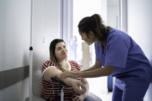 Na imagem colorida, duas pessoas estão posicionadas no centro. Uma está sentada enquanto a outra coloca medicamento no braço dela