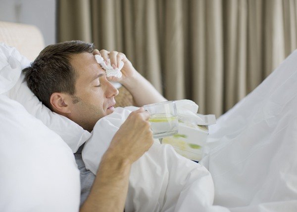 Na imagem colorida, um homem está deitado sobre uma cama. Ele está coberto por uma manta, está com uma das mãos na cabeça enquanto segura um lenço