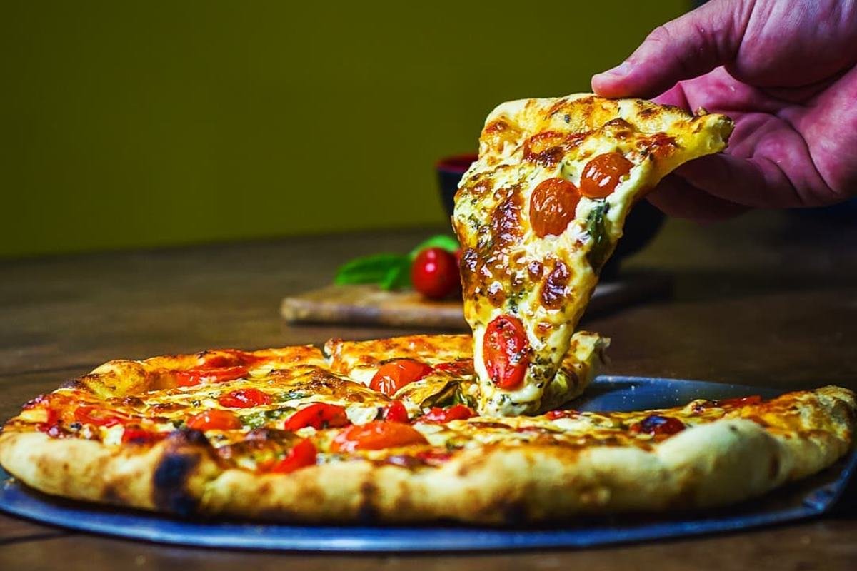 Na foto temos uma pizza e uma mão tirando uma fatia dela