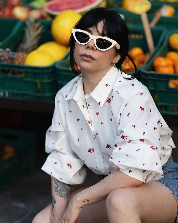 Modelo branca e com tatuagens com camisa branda da Levi's, óculos escuro. A mulher está sentada em uma caixa na feira, com frutas atrás