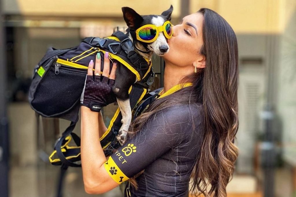 Na foto temos uma mulher vestida com roupa de ciclismo segurando uma cachorra com oculos de proteção
