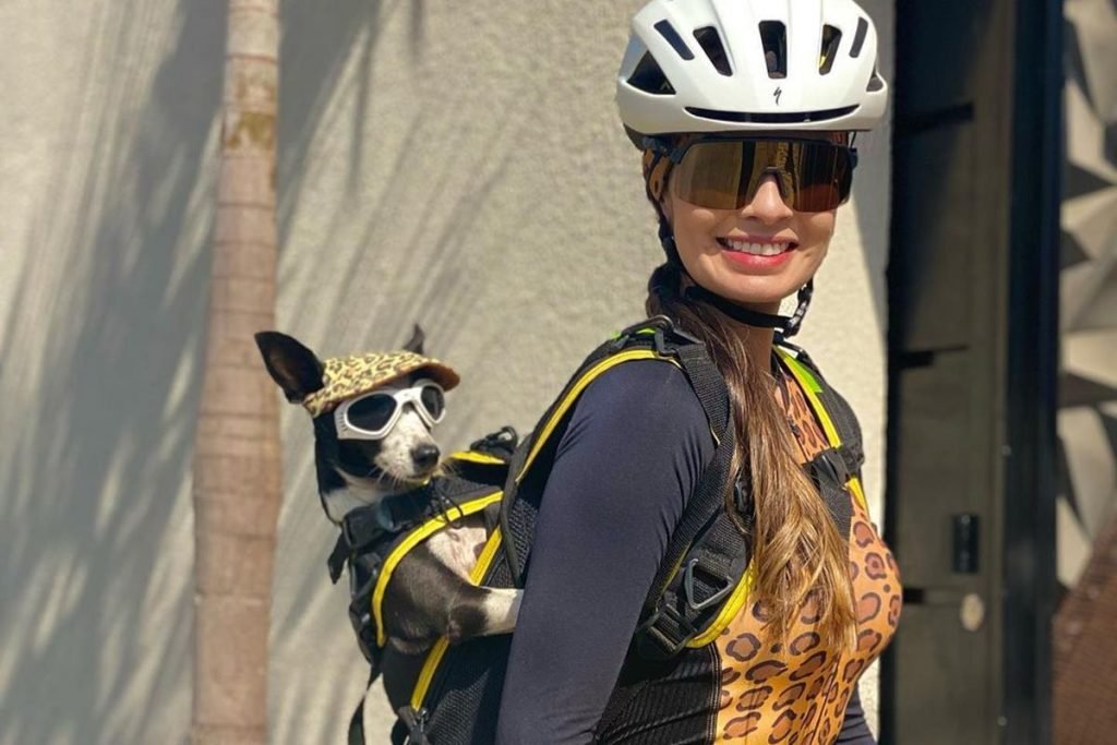 Na foto temos uma mulher com roupa, oculos e capacete de ciclismo com uma mochila e dentro uma cachorra de bone e oculos