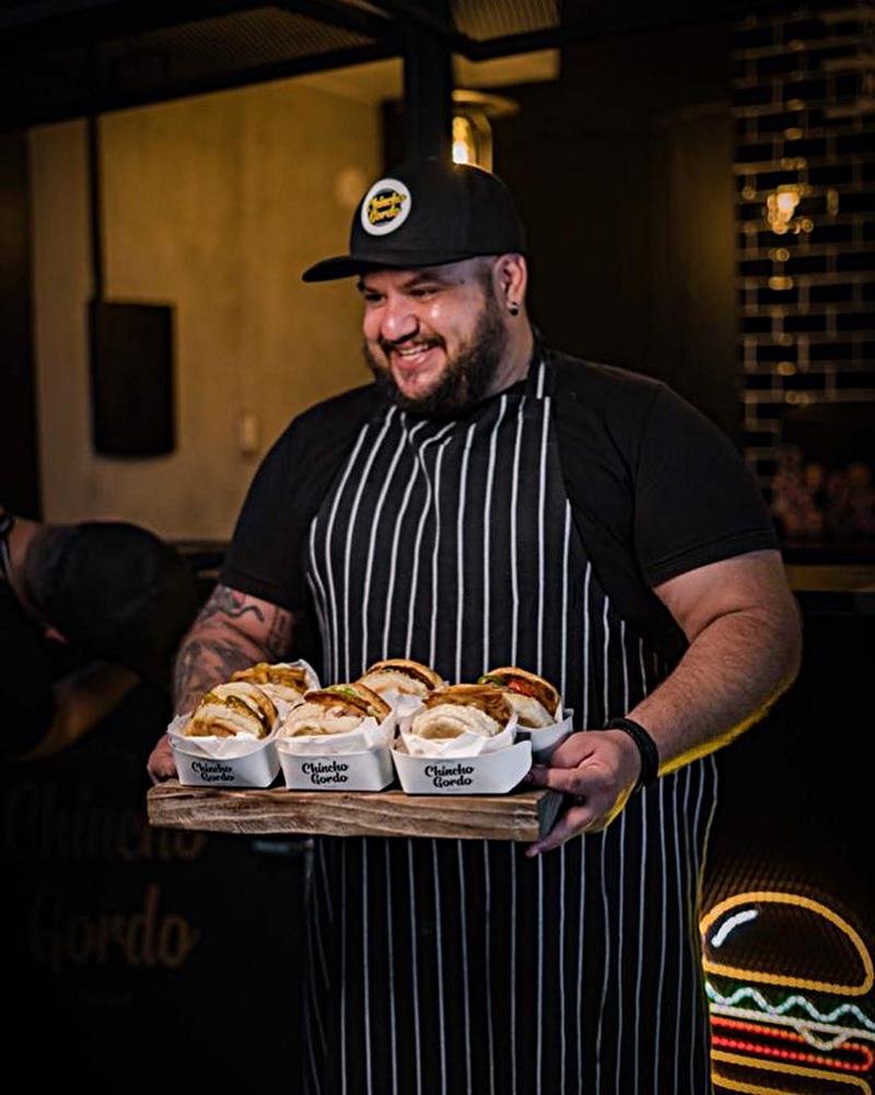Na foto, vemos um homem de avental e bone pretos com uma bandeja de hamburgueres - Metrópoles