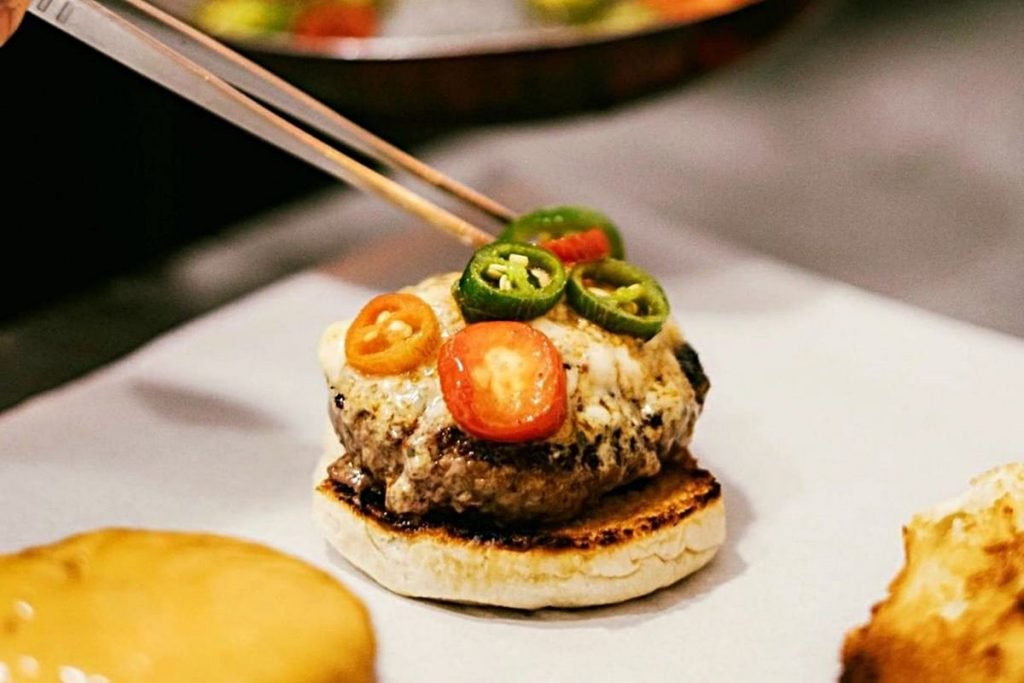 Na foto, temos um hamburguer e uma pinça colocando rodelas de pimentas - Metrópoles
