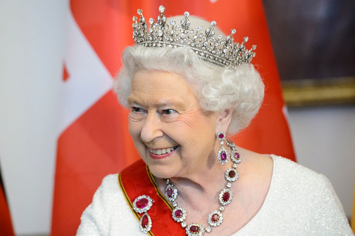 Mulher de cabelos brancos com uma coroa, joias e uma faixa vermelha
