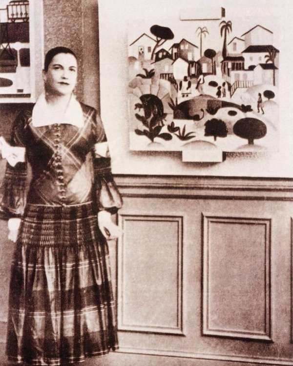 A pintora Tarsila do Amaral com vestido xadrez e um de seus quadros ao fundo
