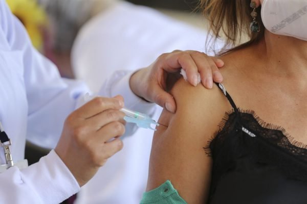 Mão aplicando vacina em braço de mulher com blusa preta de alça