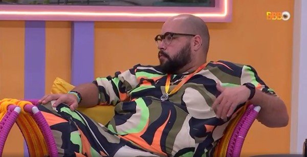 O cantor Tiago Abravanel no BBB, sentado em uma poltrona, com roupa estampada