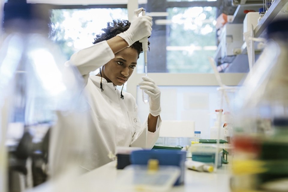 Foto colorida de mulher vestida com roupa branca em um laboratório manuseando tubos de ensaio. Foto ilustra reportagem sobre redução de orçamento para pesquisa no Brasil. / Metrópoles