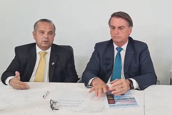 O ministro do Desenvolvimento Regional, Rogério Marinho, aparece ao lado do presidente Bolsonaro em meio a fala. Ambos possuem microfones a frente, sentados numa mesa branca, com folhas de papel espalhadas - Metrópoles