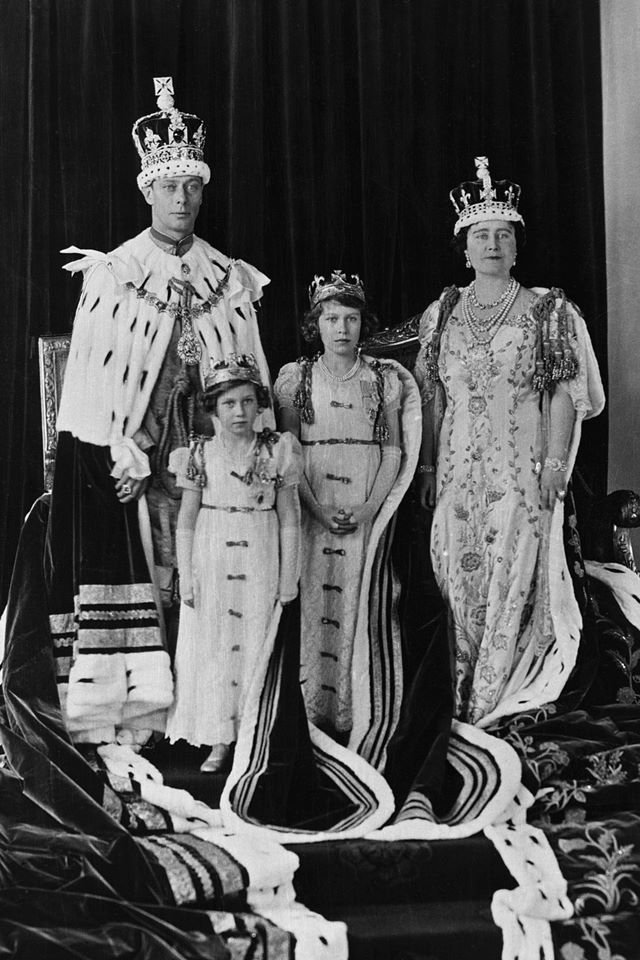 Foto em preto e branco de homem e mulher com duas meninas. Todos usam trajes e ícones da realeza