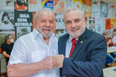 Ex-presidente Lula e o senador Jean Paul Prates, ambos do PT, posam para foto. Eles estão de frente, usam trajes sociais, e encostam os punhos num cumprimento. Ao fundo, vários cartazes de propaganda de Lula e do PT - Metrópoles