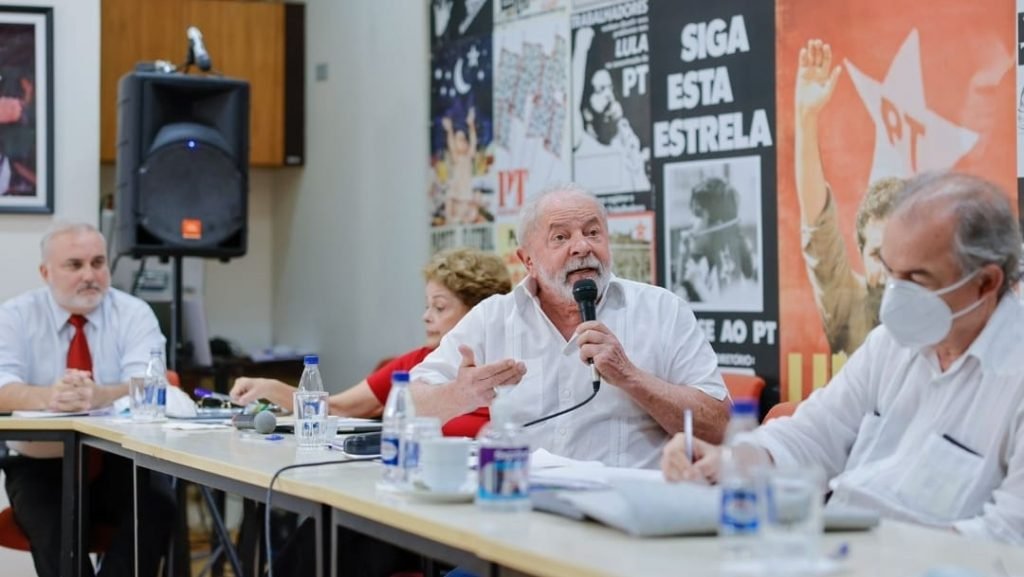 Na foto, sentados numa mesa longa, estão o senador Jean Paul Prates, a ex-presidente Dilma Rousseff, o ex-presidente Lula e o ex-senador Aloísio Mercadante, todos do PT. Lula discursa num microfone, olhando para frente, numa sala com cartazes do PT - Metrópoles