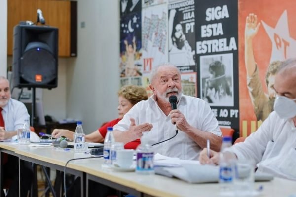 Na foto, sentados numa mesa longa, estão o senador Jean Paul Prates, a ex-presidente Dilma Rousseff, o ex-presidente Lula e o ex-senador Aloísio Mercadante, todos do PT. Lula discursa num microfone, olhando para frente, numa sala com cartazes do PT - Metrópoles